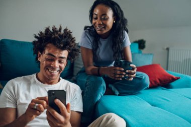 Neşeli Afrikalı Amerikalı çift hafta sonları oturma odasında oturup sohbet ederken akıllı telefon kullanıyor..