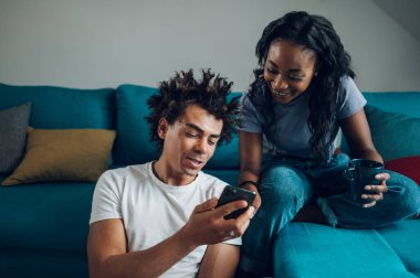 Neşeli Afrikalı Amerikalı çift hafta sonları oturma odasında oturup sohbet ederken akıllı telefon kullanıyor..