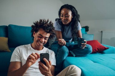 Afro-Amerikalı çift rahat evlerinde akıllı telefondan ilginç videolar izleyip boş zamanlarında kahve içiyorlar. Teknoloji ve yaşam tarzı kavramı.