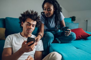 Kahve ya da çay içerken ve evde akıllı telefon kullanırken eğlenen Afro-Amerikan bir çiftin portresi. Sosyal ağlara göz atmak ya da kahve içerken video izlemek.