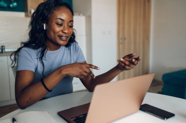 Genç Afro-Amerikalı bir kadın evdeki bir mutfakta dizüstü bilgisayarla video görüşmesi yapıyor ve iş arkadaşlarıyla internet üzerinden konferans yapıyor. Sosyal uzaklık ve modern teknoloji.