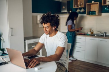 Memnuniyetle, evde bir mutfakta otururken dizüstü bilgisayarla çalışan Afro-Amerikan bir adam. Başarı işi ve evde serbest çalışma.