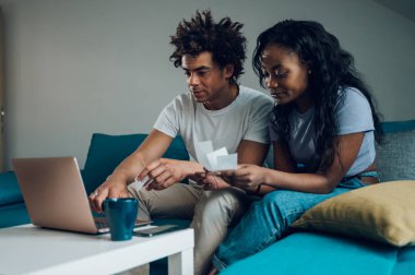 Evde dizüstü bilgisayarı ve kredi kartıyla oturan ve aylık faturaları ya da online alışverişi ödeyen Afro-Amerikan çift. Çevrimiçi bankacılık başvurusu, vergileri gözden geçirme, iç faturalar.