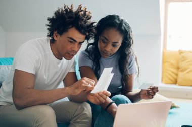 Genç Afro-Amerikan çift evde harcamalarını ve ev bütçelerini hesaplarken dizüstü bilgisayar ve kredi kartı kullanıyor. İnternetten alışveriş. Muhasebeyi ve faturaları kontrol ediyorum..