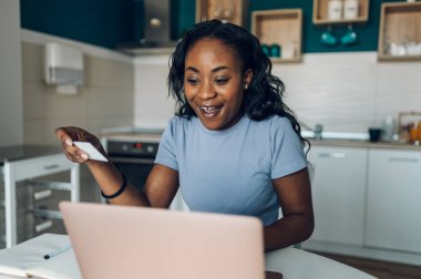 Gülümseyen Afro-Amerikalı genç bir kadın evdeki mutfakta bilgisayar ve kredi kartı kullanırken online alışveriş yapıyor. Aktarım yapılıyor.