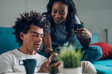 Neşeli Afrikalı Amerikalı çift hafta sonları oturma odasında oturup sohbet ederken akıllı telefon kullanıyor..