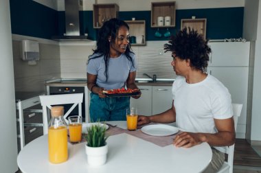 Mutlu Afro-Amerikan çifti sağlıklı yemek yerken ve portakal suyu içerken mutfakta eğleniyor. Sabahları birlikte vakit geçirmenin tadını çıkarıyorlar..
