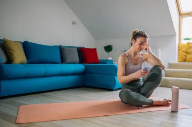 Antrenmandan sonra dinlenen ve akıllı telefon kullanan bir kadın. Evde yoga yaptıktan sonra genç ve sportif bir kadın. Yoga minderinde dinlenmek, cep telefonunda mesajlaşmak ve sakinleştirici müzik dinlemek..