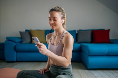 Atletik genç bir kadın, evdeki antrenmandan sonra en sevdiği müziği dinliyor ve fitness uygulamasını ya da sosyal ağları akıllı telefondan kontrol ediyor. Spor, teknoloji ve sağlıklı yaşam tarzı konsepti.