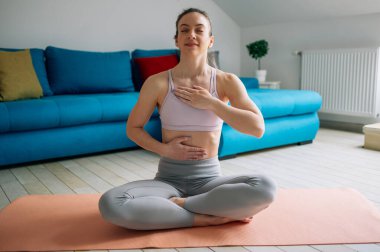 Spor bluzlu ve taytlı genç sağlıklı bir kadın evde yoga yaparken yoga minderinde oturup meditasyon yapıyor. Farkındalık meditasyon kavramı.
