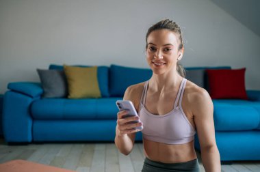 Atletik genç bir kadın, evdeki antrenmandan sonra en sevdiği müziği dinliyor ve fitness uygulamasını ya da sosyal ağları akıllı telefondan kontrol ediyor. Spor, teknoloji ve sağlıklı yaşam tarzı konsepti.