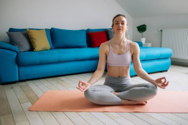 Çekici genç bir kadın evinde, oturma odasında, nilüfer yogası pozisyonunda, spor minderinin üzerinde oturuyor. Rahatlatıcı ruh hali, meditasyon. Stres yok.