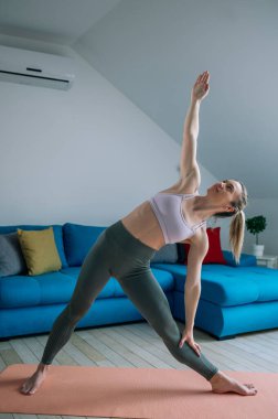 Genç atletik çekici kadın evde yoga yapıyor ve Utthita Trikonasana egzersizi yapıyor. Genişletilmiş üçgen pozu. Evde yoga..