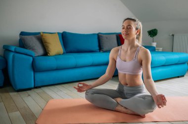 Spor bluzlu ve taytlı genç sağlıklı bir kadın evde yoga yaparken yoga minderinde oturup meditasyon yapıyor. Farkındalık meditasyon kavramı.