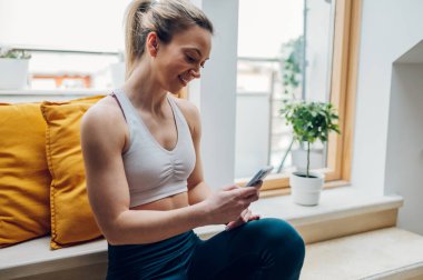 Spor yapan genç bir kadın antrenmandan sonra dinleniyor ve pencere kenarında otururken akıllı telefon kullanıyor. Egzersiz yaparken girin, bir ileti yazın.