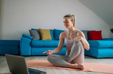 Güzel bir genç kadın evde antrenman yapıyor ve oturma odasında dizüstü bilgisayar başında yoga minderinde vücut esnetiyor. Evde egzersiz yap..