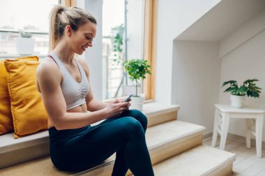Spor yapan genç bir kadın antrenmandan sonra dinleniyor ve pencere kenarında otururken akıllı telefon kullanıyor. Egzersiz yaparken girin, bir ileti yazın.