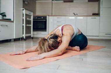 Atletik güzel bir kadın, evde bir spor paspasının üzerinde otururken, evde meditasyon için yoga çocuk pozu veriyor. Evde sağlık ve yoga..