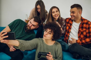 Heyecanlı bir grup arkadaş canlısı genç insan gülümseyip komik suratlar yaparken akıllı bir telefonla selfie çekiyorlar ve kanepede ve yerde oturuyorlar..