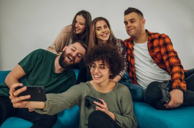 Heyecanlı bir grup arkadaş canlısı genç insan gülümseyip komik suratlar yaparken akıllı bir telefonla selfie çekiyorlar ve kanepede ve yerde oturuyorlar..