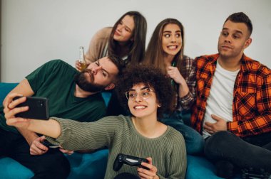 Neşeli bir arkadaş grubu evde eğleniyor ve kanepede otururken akıllı bir telefonla selfie çekiyor. Arkadaşlık ve teknoloji kavramı.