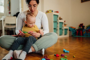 Gülümseyen bebek bakıcısı ya da dadı evde bir çocukla yerde oturup kille oynuyorlar. Anne çocuğa renkleri ve farklı şekilleri nasıl şekillendireceğini öğretir. Uzayı kopyala Çocuk gelişimi.