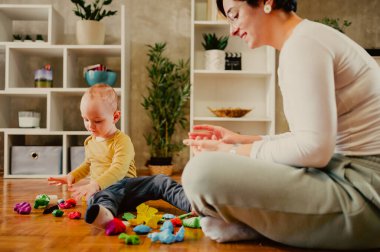 Anne ve küçük oğlu evde renkli kille oynuyor ve birlikte resim yapıyorlar. Bebek bakıcısı kadın ya da teyzesi, eğlenirken çocuğa yardım ediyor. Mutlu bir aile. Toprağı şekillendiren bir çocuğa odaklan..