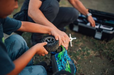Uzaktan kumandalı arabaları tamir ederken çekilmiş bir resim. Uzaktan kumandalı oyuncak bir arabanın lastiğini tamir eden çocuk ve adamın elleri. Babası alet çantası kullanırken çocuk lastik değiştiriyor..