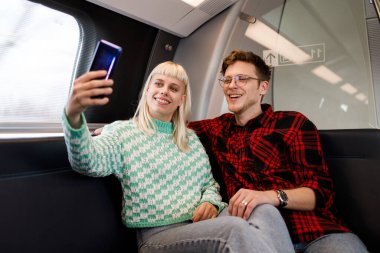 Genç bir çift metroya biniyor ve selfie çekiyor. Metro treninde eğlenen ve telefonda gülümseyen bir çiftin portresi. Umumi trenle seyahat edenler