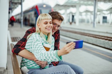 Tren istasyonunda oturan genç hippi yolcuların portresi. Telefona gülümserken sarılıp selfie çekiyorlar. Tren istasyonunda tren bekleyenler ve kendi portrelerini çekenler..