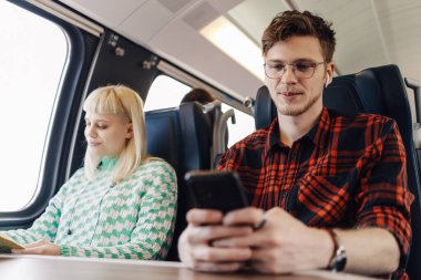 Trende oturan genç bir gezginin portresi. Kulağında kulaklıklar ve telefonda gülümseyen bir şekilde telefonla mesajlar yazıyor. Yanında kitap okuyan genç bir kadın var..