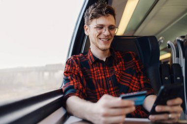 Tran 'da pencerenin yanında oturan ve kredi kartı ve akıllı telefon kullanarak internetten alışveriş yapan trenin portresi. Genç bir trend yolcusu toplu taşıma ve online alışveriş ile gidip geliyor