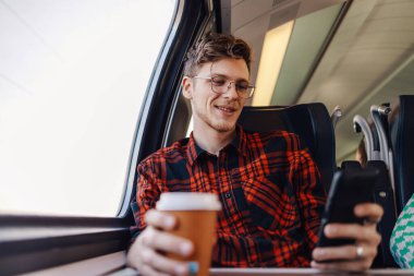 Mutlu bir gezgin trende oturuyor ve kahve içerken telefonda mesajlar yazıyor. Genç bir yolcu, toplu taşıma araçlarıyla seyahat ederken telefona gülümsemek için kahve içiyor.