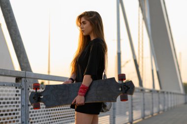 Genç bir kadın uzun tahtasını tutuyor ve köprüde duruyor.