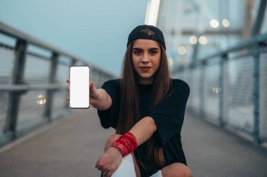 Genç bir kadın uzun tahtasının üzerinde oturuyor ve köprüdeyken akıllı telefon gösteriyor.