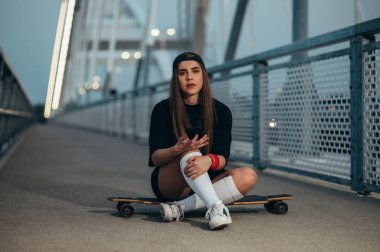 Genç bir kadın uzun tahtasının üzerinde oturuyor ve köprüdeyken akıllı telefon kullanıyor.