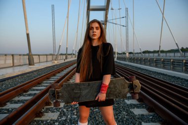 Genç bir kadın uzun tahtasını tutuyor ve köprüde duruyor.