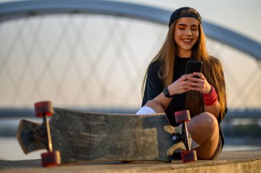 Genç bir kadın nehir kenarında oturuyor ve uzun tahta üstüne akıllı telefon kullanıyor.