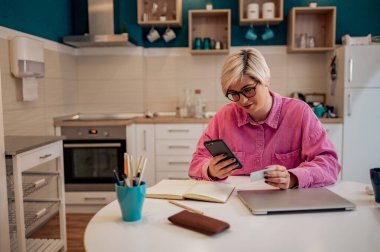 Genç beyaz kadın tüketici elinde kredi kartı ve akıllı telefon tutarken mutfaktaki masada oturup online bankacılık işlemlerini yapıyor. Çevrimiçi sanal alışveriş, güvenli mobil bankacılık.
