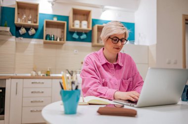 Genç, güzel, sarışın, kısa saçlı bir kadın evinden uzak bir şirkette çalışıyor. Mutfakta dizüstü bilgisayarı var ve serbest çalışıyor. Evde çalışıyorum. Çevrimiçi iş ve serbest çalışan.