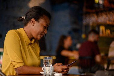 Güzel, Afro-Amerikalı bir kadın akıllı telefon kullanıyor ve kafede otururken kahve içiyor.