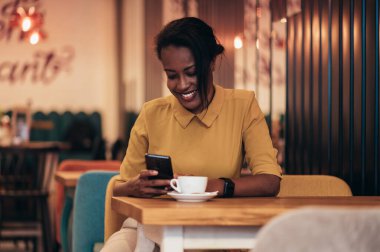 Güzel, Afro-Amerikalı bir kadın akıllı telefon kullanıyor ve kafede otururken kahve içiyor.