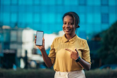 Güzel, genç, Afro-Amerikalı bir kadın. Şehir caddesinde dikilirken akıllı telefon gösteriyor.