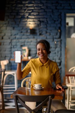 Beautiful young african american woman showing a smartphone while sitting in a cafe clipart