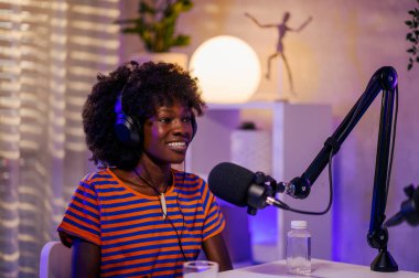 Irklar arası bir podcast sunucusunun portresi evde kayıt ve yayın stüdyosunda oturuyor ve kulaklık takıp mikrofona gülümserken bir podcast bölümü çekiyor. İçerik oluşturucu bir podcast yapıyor