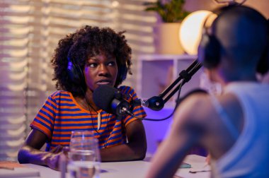 Düşünceli, çok ırklı bir konuk kulaklık takıyor ve mikrofonda bir stüdyoda podcast sunucusuyla otururken ciddi konular hakkında konuşuyor. İki kız canlı yayında.