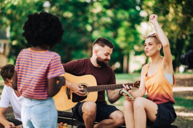 Genç bir sokak müzisyeni parkta çok kültürlü gençler için gitar çalıyor. Yetenekli bir sokak müzisyeni şehir parkında gitar çalıyor. Etrafındaki gençler dinleyip dans ediyorlar.