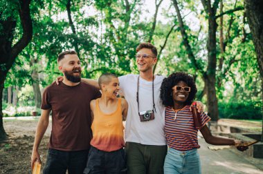 Irklar arası en iyi arkadaşlar bir şehir parkında sarılıp yürüyorlar. Güneşli bir yaz gününde şehir parkında yürürken kucaklaşan ve gülümseyen çok kültürlü küçük bir grubun portresi..