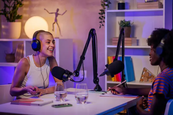 stock image Two cheerful journalists are sitting in a home podcasting studio with headphones on and speaking at the microphone during the podcast. Two multiracial podcasters are going live on air and streaming.