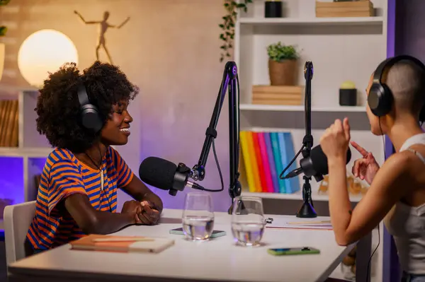 stock image Two multiracial cheerful content creators are talking about fun topics on a podcast while sitting in a home broadcasting studio. Two gen z girls are discussing fun topics in a home recording studio.